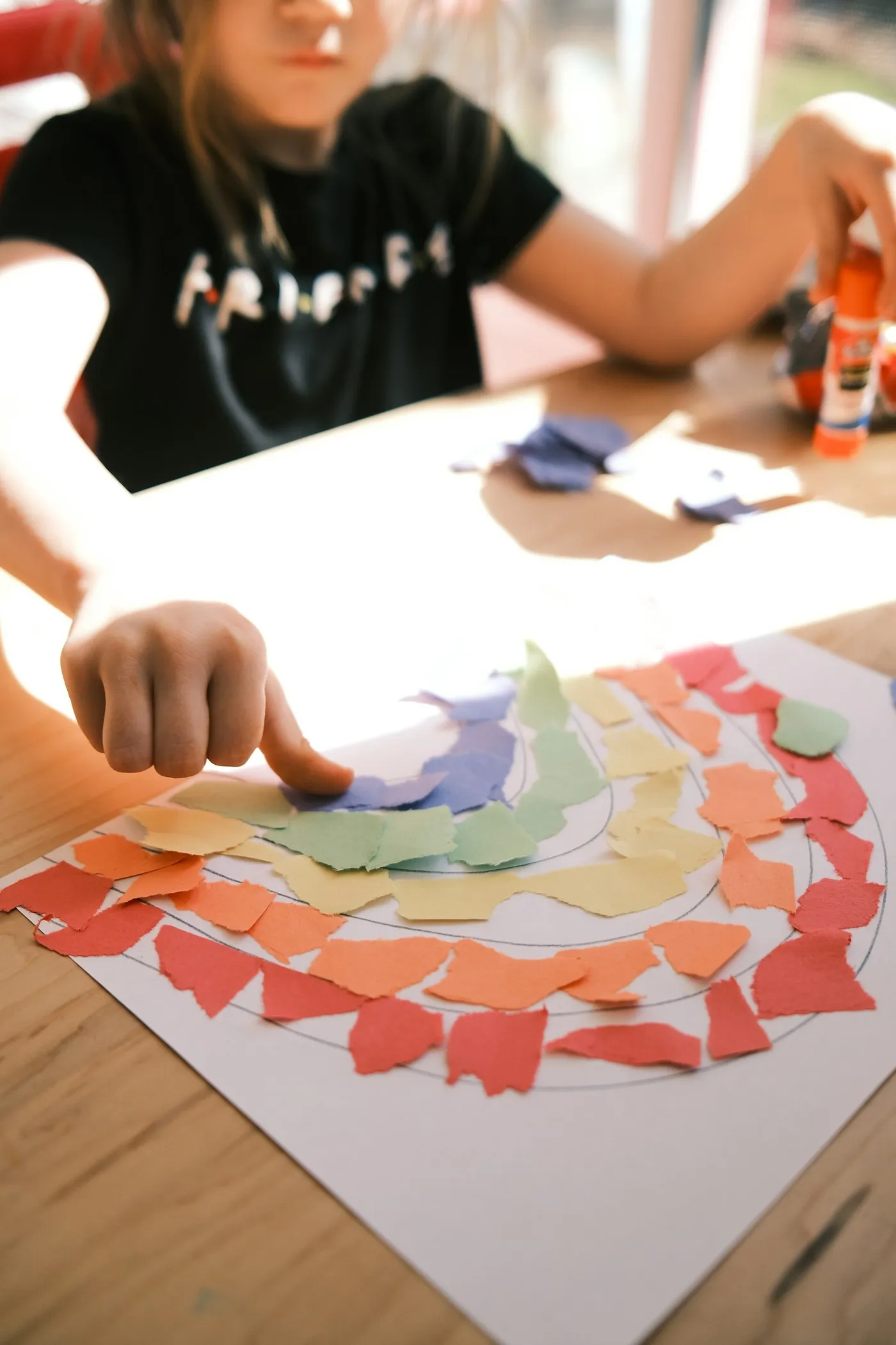 Una niña pega trozos de papel de colores sobre el dibujo de un arcoíris.
